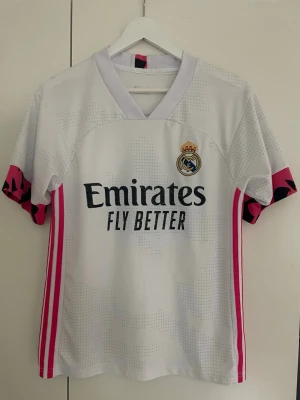 Real Madrid matchtröja Benzema #9 - Säljer en vit Real Madrid matchtröja med rosa detaljer och svarta tryck. Tröjan har klubbmärke, Emirates Fly Better på bröstet och Benzema samt nummer 9 på ryggen. Korta ärmar med rosa och svarta mönster på mudden. Perfekt för fotbollsfans!