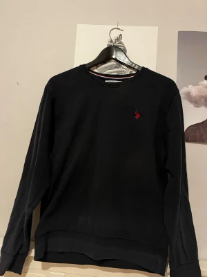 U.S. Polo Assn. - En svart sweatshirt med rund halsringning och en liten röd broderad logga på bröstet. Tröjan har långa ärmar och ribbade muddar vid ärmslut och nederkant. Perfekt för en avslappnad och stilren look.