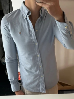 Ljusblå skjorta från Ralph Lauren - Klassisk ljusblå skjorta från Ralph Lauren med button-down krage och broderad logga på bröstet. Skjortan har långa ärmar, normal passform och är tillverkad i mjuk bomull. Perfekt till jeans eller chinos för en clean look.