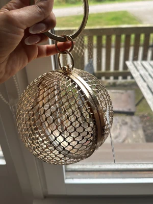 Guldig rund mesh handväska - Unik handväska i form av en rund boll med mesh-design i guldigt metallmaterial. Väskan har ett stort ringhandtag och öppnas med ett gångjärn på mitten. Perfekt för dig som vill sticka ut med accessoarer som verkligen syns.