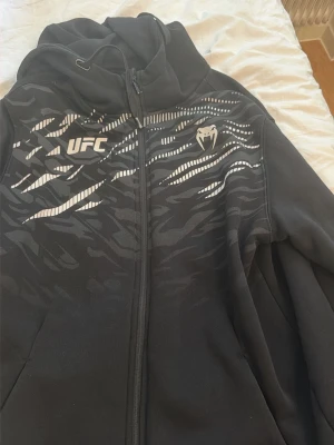 Svart UFC hoodie från Venum - Säljer en svart hoodie från Venum med UFC-logga på bröstet och coolt mönster i vitt och grått över axlar och bröst. Dragkedja framtill, huva med snören och mjukt material. Perfekt för dig som gillar sportig stil och vill sticka ut.