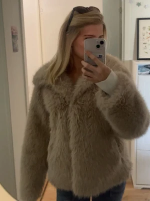 Beige fluffig pälsjacka - Supersnygg beige pälsjacka med oversized passform och bred krage. Jackan är gjord i fuskpäls som känns mjuk och lyxig. Perfekt för att hålla värmen under kalla dagar och ger en trendig look till alla outfits.