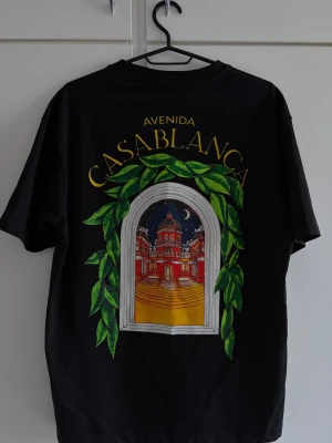 Svart Avenida Casablanca t-shirt - Svart t-shirt från Casablanca med färgglad grafisk print på ryggen som föreställer en fönsterutsikt mot en byggnad och natthimmel, omgiven av gröna blad. Texten 'Avenida Casablanca' i gult ovanför motivet. Klassisk passform och rund hals.