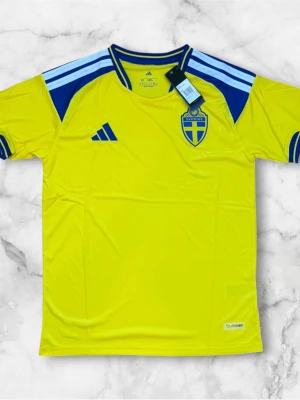 Sverige matchtröja Adidas gul/blå - Säljer en officiell Sverige matchtröja från Adidas i gul med blå detaljer och blått landslagsemblem på bröstet. Tröjan har korta ärmar, rund hals och klassiska Adidas-ränder på axlarna. Tillverkad i lätt och ventilerande funktionsmaterial.
