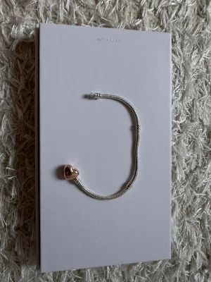 Pandora armband med hjärtlås - Snyggt armband från Pandora i silverfärg med ett roséguldigt hjärtlås. Kedjan är tunn och elegant, perfekt för att kombinera med andra smycken eller bära som det är. Passar dig som gillar stilrena detaljer och klassisk design.