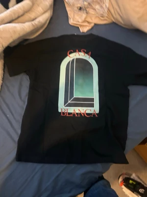 Svart Casablanca t-shirt med tryck - Snygg svart t-shirt från Casablanca med stort grafiskt tryck i turkos, vitt och rött på ryggen och mindre tryck framtill. Klassisk passform och rund hals. Perfekt för dig som gillar streetwear och vill sticka ut med en unik design.