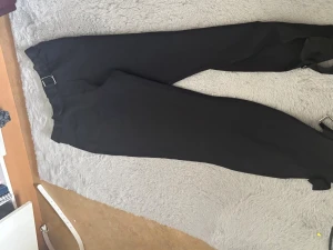 Svarta cargobyxor med spännen - Säljer ett par svarta cargobyxor med coola justerbara spännen i midjan och vid bensluten. Byxorna har en rak passform och är tillverkade i ett slitstarkt tyg som ger en streetwear-vibe. Perfekta för dig som gillar en edgy och avslappnad stil. Helt oanvänd, med prislappen.