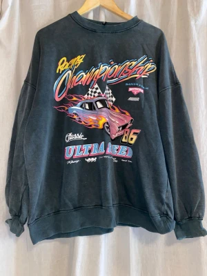 Oversized racing sweatshirt med tryck - Cool oversized sweatshirt i mörkgrått med färgglatt racing-inspirerat tryck framtill. Motiv med bil, eldflammor och text som 'Racing Championship' och 'Ultra Speed'. Tröjan har rund hals, ribbade muddar och avslappnad passform. Perfekt för dig som gillar streetwear och retrostil.