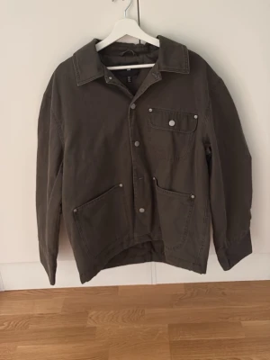 Olivgrön overshirt från H&M - Stilren olivgrön overshirt från H&M med tre fickor framtill och tryckknappar. Overshirten har klassisk krage och är gjord i ett slitstarkt bomullstyg. Perfekt att bära öppen över en t-shirt för en avslappnad look.