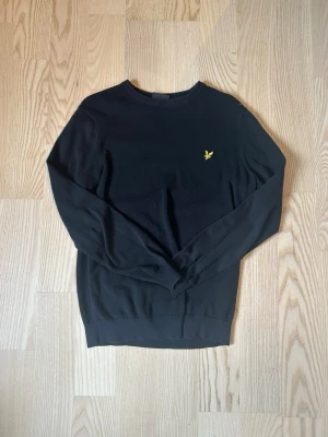 Svart tröja från Lyle & Scott - Klassisk svart tröja från Lyle & Scott med rund halsringning och den ikoniska gula örnloggan broderad på bröstet. Tröjan har långa ärmar och ribbade muddar vid ärmslut och nederkant. Perfekt till en stilren och enkel look.