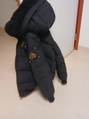 Svart dunjacka från Moose Knuckles - Snygg svart dunjacka från Moose Knuckles med guldfärgade dragkedjor och detaljer. Jackan har flera fickor med dragkedja och en stor, fluffig huva med svart fuskpäls. Perfekt för kalla vinterdagar och riktigt varm. Har kvittot också 