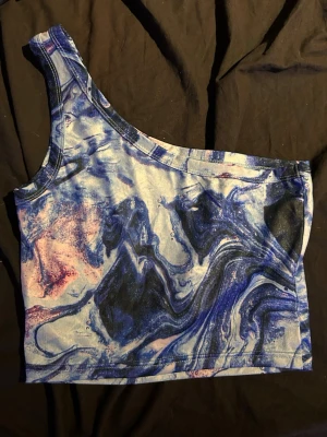Blå oneshoulder croppad topp - Säljer en croppad oneshoulder topp med blått och lila marmormönster. Toppen är ärmlös och har en cool asymmetrisk design 