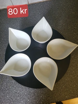 Snurrbricka  - Säljer en snurrbricka med fem vita skålar i droppform, perfekta för snacks, såser eller småplock. Skålarna är tillverkade i porslin och har en modern, stilren design som passar in i de flesta kök. Snygga att ställa fram på bordet när du har gäster. Den lila skålen i mitten saknas dock. Och den har tappat lite färg som man kan se på bilden.