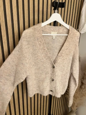 Kofta i alpackaull blend från H&M - Mysig beige kofta från H&M med v-ringning och tre stora knappar framtill. Koftan är stickad i ett mjukt material och har en avslappnad, något croppad passform med långa ärmar och ribbade muddar.