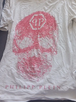 Vit Philipp Plein t-shirt med rosa tryck - Snygg vit t-shirt från Philipp Plein med stort rosa grafiskt dödskalletryck på framsidan och logga. Klassisk passform och korta ärmar. Tillverkad i mjuk bomull som känns skön mot huden. Perfekt för dig som gillar statement-plagg och streetstyle.