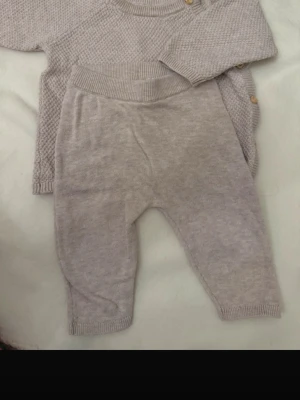 Stickat set i ljusbeige med knappar - Mysigt stickat set i ljusbeige med tröja och byxor. Tröjan har rund halsringning, ribbade muddar och dekorativa träknappar längs sidan. Byxorna har bred resår i midjan och ribbade benslut. Perfekt för en avslappnad och stilren look.