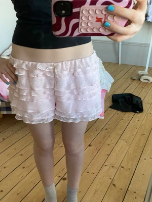 Rosa volangpyjamasshorts från Monki - Supersöta ljusrosa pyjamasshorts från Monki med flera lager volanger och liten rosett framtill. Tillverkade i ett mjukt, ribbat material som känns skönt mot huden. Perfekta för mysiga hemmakvällar eller sommarens varma nätter.