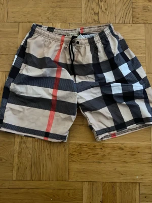 Burberry rutiga badbyxor - Snygga badbyxor från Burberry med klassiskt rutmönster i beige, svart, vitt och rött. De har resår i midjan med svart dragsko och nätfoder på insidan. Perfekta för stranden eller poolen om du vill sticka ut med stil.