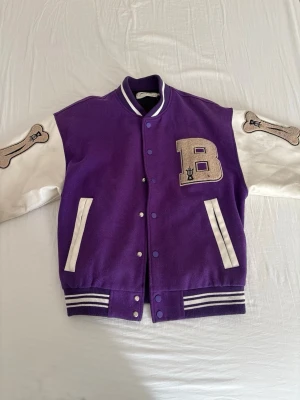 Lila bomberjacka med vita ärmar - Snygg lila bomberjacka med vita ärmar och ribbade muddar i lila och vitt. Stor bokstav 'B' i beige på bröstet och coola benpatches på ärmarna. Två fickor framtill och tryckknappar. Perfekt för dig som vill sticka ut med en retro varsity-look.