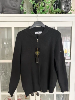 Svart ziptröja från Stone Island - Snygg svart ziptröja från Stone Island i ribbstickad bomull. Tröjan har hel dragkedja framtill, rundad krage och den klassiska Stone Island-loggan på taggen. Perfekt för dig som gillar stilrena och tidlösa plagg.