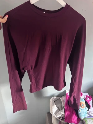Vinröd långärmad tröja - En enkel och stilren vinröd långärmad tröja med rund halsringning. Tröjan är rak i modellen och tillverkad i ett mjukt, tunt bomullsliknande material. Perfekt att matcha med jeans eller kjol för en avslappnad look.