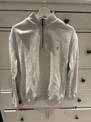 Grå halvzip från Polo Ralph Lauren - Snygg ljusgrå halvzip-tröja från Polo Ralph Lauren med broderad logga på bröstet. Tröjan har hög krage, dragkedja framtill och är tillverkad i mjukt bomullsmaterial. Perfekt för lager på lager och enkel att matcha med jeans eller chinos.