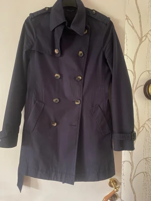 Mörkblå trenchcoat från Tommy Hilfiger - Stilren mörkblå trenchcoat med dubbelknäppning, axelklaffar och bälte i midjan. Klassisk krage och fickor på sidorna. Justerbara remmar vid ärmsluten för extra detalj. Perfekt för dig som vill ha en tidlös och snygg jacka till hösten.