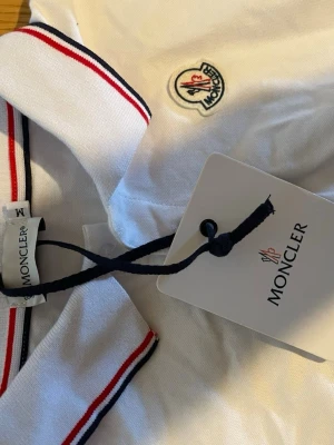 Vit Moncler pikétopp med rödblå detaljer - Säljer en vit pikétopp från Moncler med klassisk krage och knappar framtill. Kragen har snygga rödblå ränder och bröstet pryds av Monclers ikoniska logga. Perfekt för dig som gillar stilrena och sportiga plagg.
