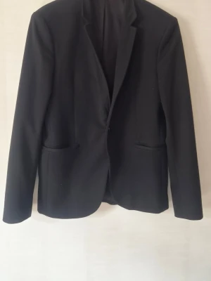 Svart Zara klassisk kavaj - En stilren svart kavaj med enkelknäppning och två framfickor. Kavajen har smal passform och klassisk krage. Perfekt för en clean och snygg look. Materialet ser ut att vara en mjuk och slät textil, troligen polyesterblandning.