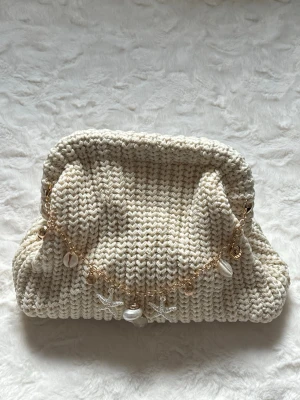 Stickad beige kuvertväska med snäckor - Superfin stickad kuvertväska i beige med chunky struktur. Väskan har en guldfärgad kedja dekorerad med snäckor och sjöstjärnor som ger en somrig och bohemisk vibe. Perfekt accessoar för dig som gillar strandinspirerad stil.