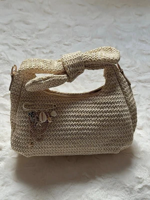 Beige virkad handväska med snäckdetaljer - Snygg beige handväska i virkat material med knut på handtaget. Väskan har dekorativa hängen med snäckor, kedjor och små berlocker. Perfekt accessoar för en trendig och avslappnad look. Ingen synlig logga eller märke.