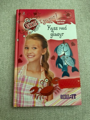 Kyss med glasy, 100% tjejer - Rolig och färgstark ungdomsbok med titeln 'Kyss med glasy' av Karen Christine Angermayer. Omslaget är rött och vitt med illustrationer av fiskar, hjärtan och en krabba. Perfekt för dig som gillar lättläst och romantisk skönlitteratur.