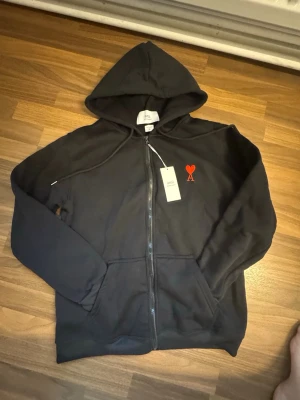 Ami Paris - Black hoodie - Säljer en svart hoodie från Ami Paris med dragkedja och huva. På bröstet finns Ami:s ikoniska röda hjärta med bokstaven A. Hoodien har fickor framtill och är gjord i mjukt bomullsmaterial. Perfekt för en chill och stilren look. Inga defekter