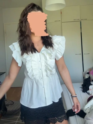 Vit blus med volanger och knappar - Snygg vit blus med volanger längs axlar och bröst, knappar framtill och v-ringning både fram och bak. Blusen har korta ärmar och är tillverkad i ett lätt tyg som ger en luftig känsla. Perfekt för att skapa en trendig och feminin look. Passar storlek S. 