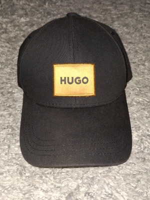 Svart keps från HUGO - Snygg svart keps från HUGO med gul patch och tydlig logga framtill. Klassisk böjd skärm och justerbart spänne bak. Tillverkad i slitstark bomull som ger en clean och modern look.
