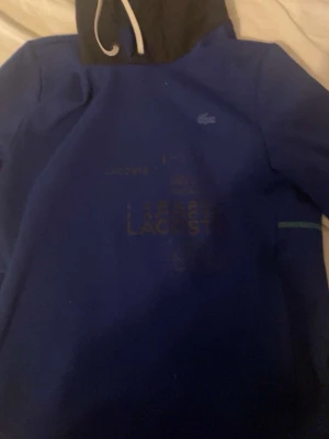 Lacoste hoodie - Mörkblå Lacoste huvtröja med svart huva och vita dragsnören. Sportig och stilren design med Lacoste logga på bröstet och diskreta detaljer framtill. Bekväm passform som passar både vardag och fritid.
