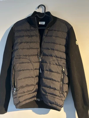 Svart pufferjacka från Moncler - Svart pufferjacka från Moncler med quiltad front och stickade ärmar. Jackan har två dragkedjeförsedda fickor, hög krage och Moncler-logga på vänster ärm. Materialmixen ger en modern och sportig känsla.