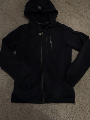 Svart zip-hoodie från SAIL RACING - Snygg mörk mörk blå hoodie med dragkedja från SAIL RACING. Modellen har huva med tryck, bröstficka med dragkedja och ribbade muddar. Perfekt för en avslappnad och sportig stil.