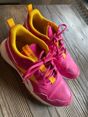 Färgglada Reebok sneakers rosa/gul - Snygga sneakers från Reebok i stark rosa färg med gula och vita detaljer. Skorna har en låg profil, klassisk snörning och en slitstark yttersula i gummi. Insidan är vadderad i gult tyg för extra komfort. Perfekta för dig som vill sticka ut!