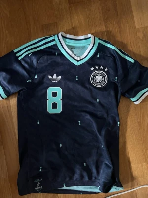 Tysklands retro fotbollströja Adidas - Mörkblå retro fotbollströja från Adidas med nummer 8 och namnet Goretzka på ryggen. Tröjan har ljusblå och vita detaljer, tre ränder på axlarna och Tysklands landslagsemblem med fyra stjärnor på bröstet. Tillverkad i lätt och ventilerande material.