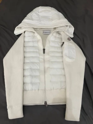 Vit hoodie från Moncler med quiltad front - Snygg vit hoodie från Moncler med quiltad front och huva. Tröjan har ribbstickade ärmar och nederkant, dragkedja framtill och en liten ficka på ärmen med Moncler-logga. Perfekt för dig som gillar exklusiv streetwear och vill ha något både stilrent och bekvämt.