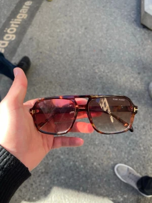 Bruna Tom Ford solglasögon - Snygga solglasögon från Tom Ford med brunskiftande bågar i klassisk pilotmodell. Glasen är lätt tonade och bågarna har guldfärgade detaljer vid gångjärnen. Perfekt accessoar för dig som vill ha en stilren och trendig look.