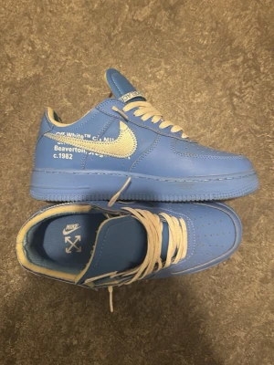 Nike Off-White Air Force 1 University Blue - Säljer ett par Nike Air Force 1 Off-White sneakers i blått läder med vita detaljer och text på sidan. Skorna har Off-White-logga på innersulan, silverfärgad Swoosh och vita skosnören. Ikoniska citattecken runt 'AIR' och röd detalj vid hälen. Använd 2-3 gånger, ej creasade och är dessutom Stock-X verifierade.