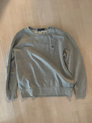 Grå sweatshirt från Polo Ralph Lauren - Klassisk grå sweatshirt från Polo Ralph Lauren med rund halsringning och diskret broderad logga på bröstet. Tröjan har ribbade muddar vid ärmslut och nederkant. Perfekt för en avslappnad och stilren look.