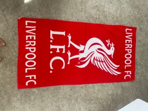 Liverpool FC röd sovpläd - Röd sovpläd med stort vitt Liverpool FC-tryck och Liverbird-logga. Perfekt för dig som älskar fotboll och vill ha något mjukt och skönt att slappa i hemma. Pläden är rektangulär och har tydliga klubbdetaljer i vitt mot den röda bakgrunden.