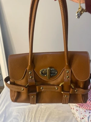 Brun handväska - Brun vintage handväska i äkta läder med dubbla handtag och guldfärgade metalldetaljer. Väskan har ett klassiskt spänne framtill, dekorativa remmar och nitar samt vit kontrastsöm. Varan är i mycket gott skick då den knappt kommit till användning. För fler frågor eller bilder är det bara att skiva! 💕