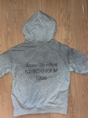Grå hoodie från Acne Studios - Säljer en grå hoodie från Acne Studios med tryck på ryggen där det står 'Acne Studios STOCKHOLM 1996'. Klassisk modell med huva och ribbade muddar vid ärmslut och nederkant. Perfekt för en avslappnad och stilren look.