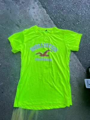 Neongul Hollister t-shirt - Säljer en neongul t-shirt från Hollister med tryck i vitt och rött framtill där det står 'Hollister California' och en ikonisk fågel. T-shirten har rund halsringning och korta ärmar. Perfekt för dig som vill sticka ut med färg!