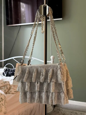 Silverfärgad axelväska med volanger Zara - Svart festväska från Zara med silverfärgad kedjerem. Väskan är täckt av små dekorativa plaststenar som ger ett glittrande och skimrande intryck när ljuset träffar dem.  Lätt och smidig storlek med plats för mobil, kort, nycklar och andra småsaker. Perfekt till middag, bröllop, student, nyår eller utekvällar.  Helt ny med prislapp kvar.