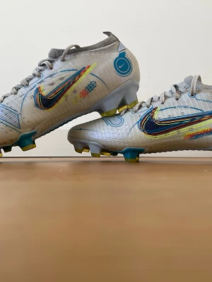 Nike Mercurial fotbollsskor vita - Säljer ett par Nike Mercurial fotbollsskor i vitt med färgglada detaljer och blått swoosh-märke. Skorna har låg profil, snörning och dobbar i gult och blått för bra grepp på planen. Tillverkade i syntetmaterial för lätt känsla och bra bollkontroll. FG dubbar.